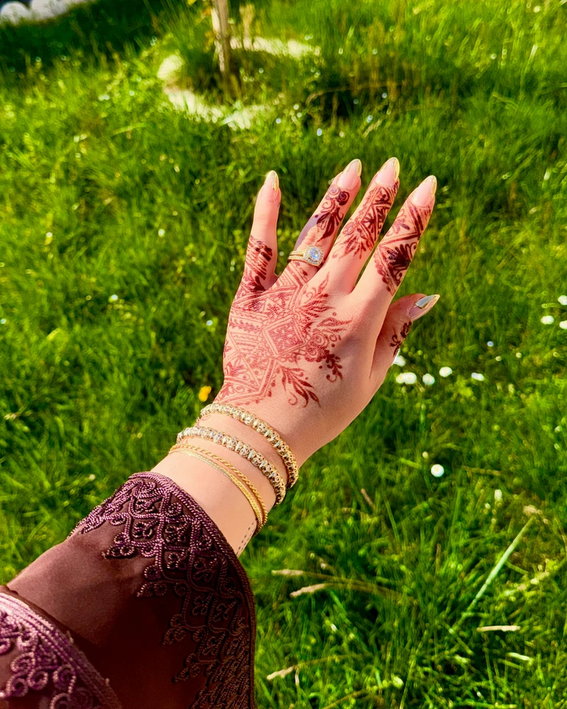 Main ornée de henné et de bracelets dorés, manche de caftan brodée, sur fond de jardin