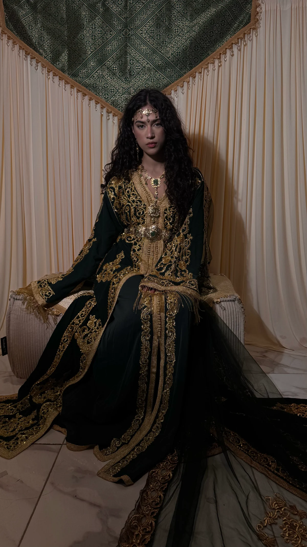Mariée en caftan vert royal brodé d’or, assise devant un rideau de soie